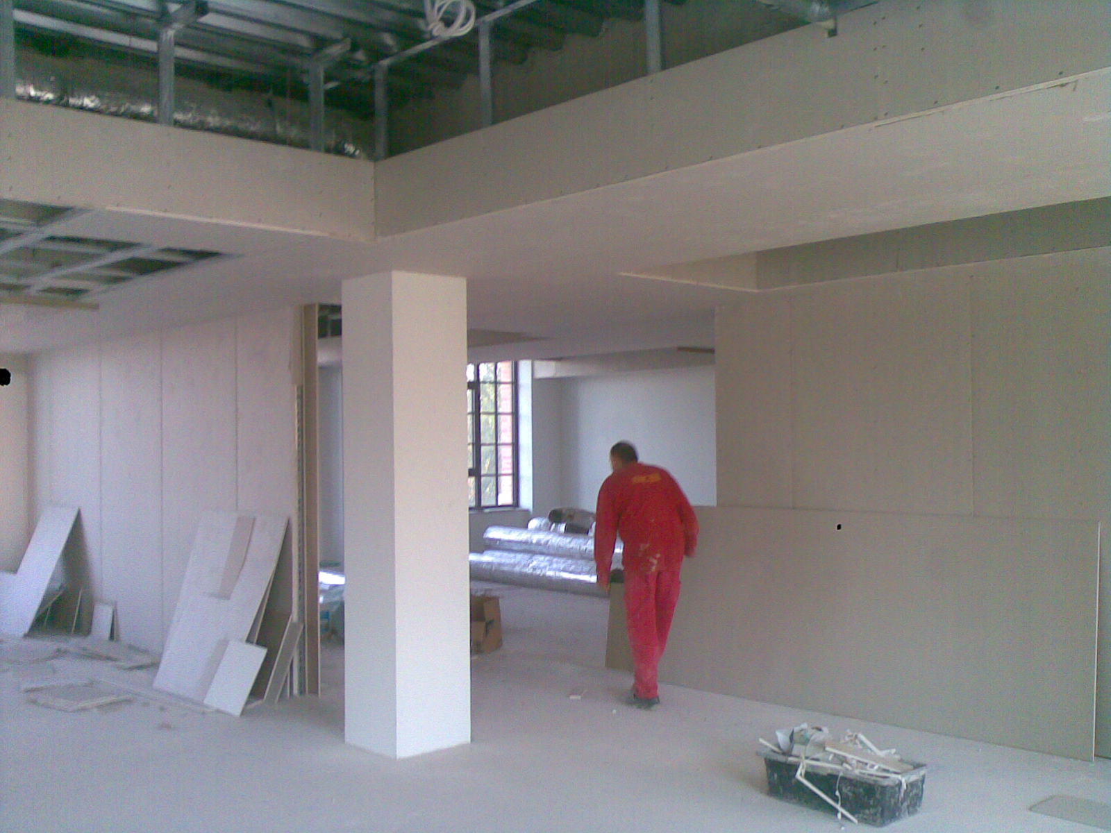 Wnętrze w trakcie montażu ścian działowych z płyt gipsowo-kartonowych, widoczny pracownik w czerwonym kombinezonie, metalowa konstrukcja sufitu podwieszanego oraz stos płyt G-K opartych o ścianę.