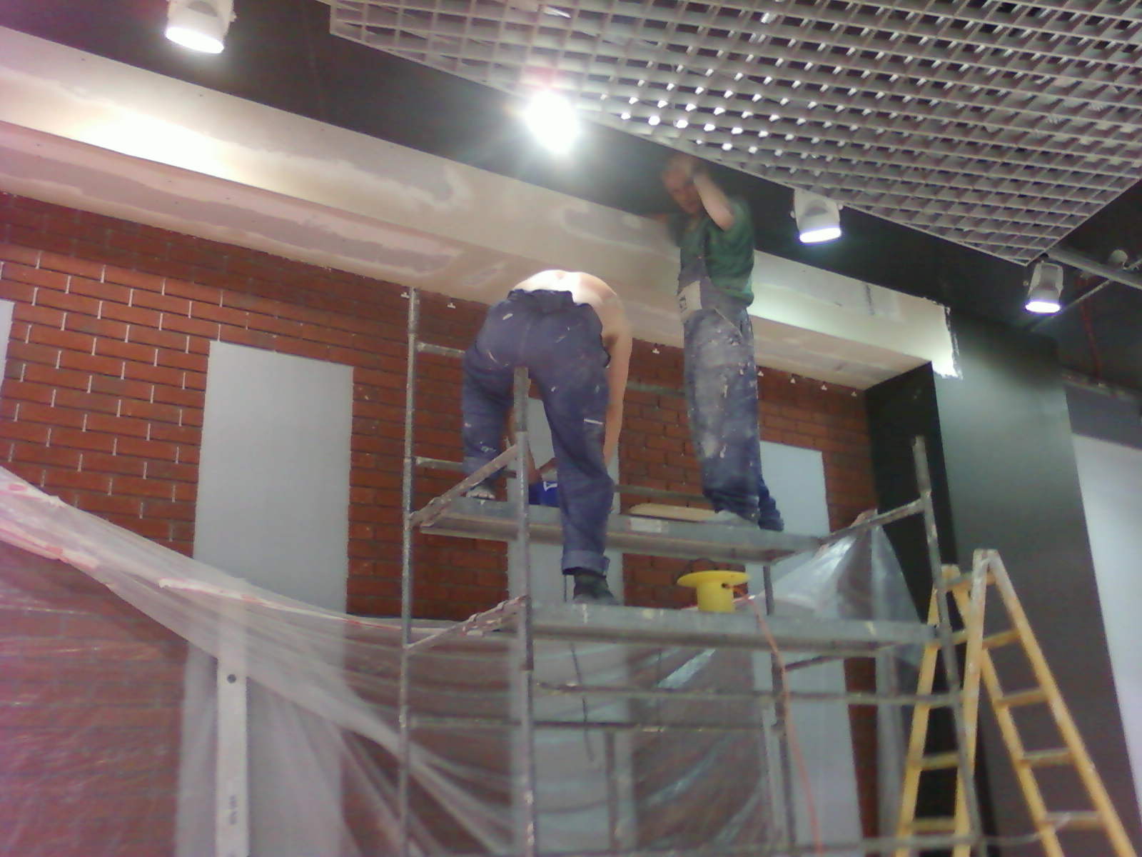 Dwóch pracowników na rusztowaniu, jeden szpachluje sufit G-K, drugi przygotowuje ścianę z cegły do dalszych prac wykończeniowych.