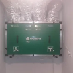 Zielony rekuperator EkoZefir zamontowany w pomieszczeniu, widoczne kanały wentylacyjne i oznaczenia 'Nawiew', 'Wywiew', 'Czerpnia', 'Wyrzutnia'.