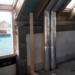 TERALWENT Tomasz Kufel - Dwa izolowane przewody wentylacyjne przechodzące przez otwory w podłodze na poddaszu w trakcie montażu, widoczne bloczki betonowe, drewniane elementy konstrukcyjne dachu i zwój niebieskiego...