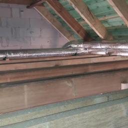 TERALWENT Tomasz Kufel - Izolowane przewody wentylacyjne w przestrzeni poddasza, widoczne drewniane elementy konstrukcyjne dachu i fragment ściany z bloczków.