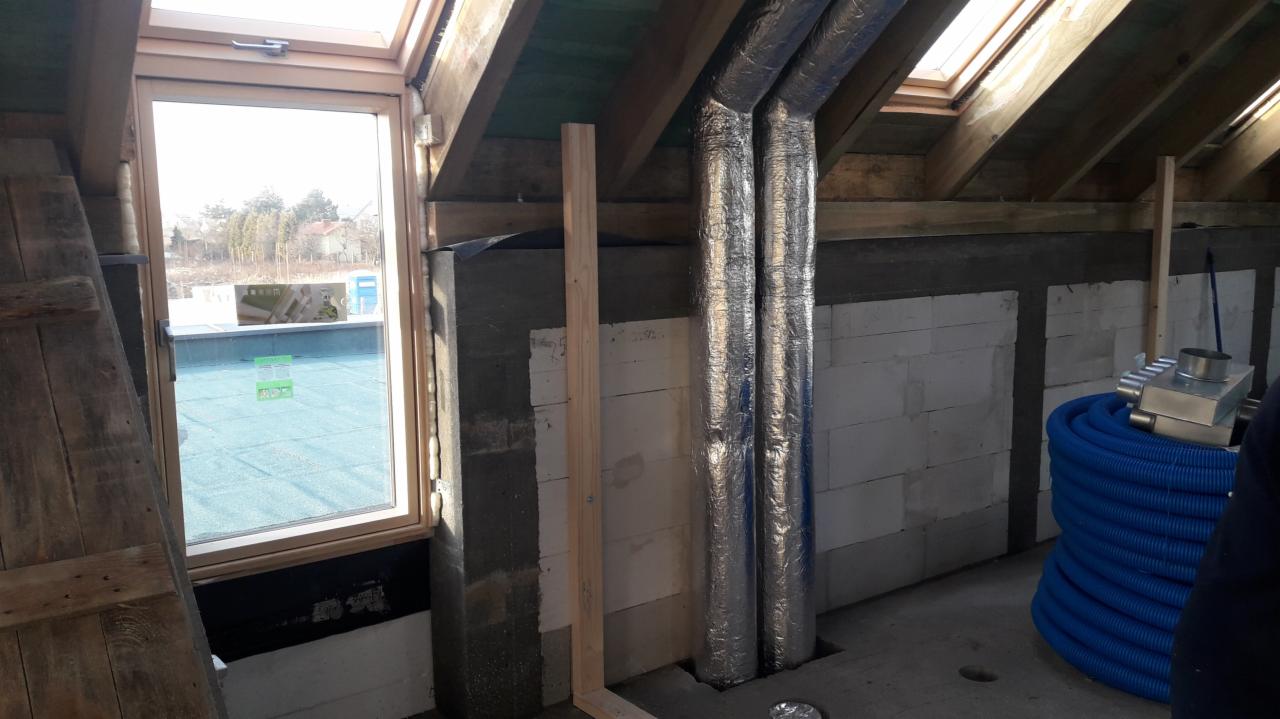 Dwa izolowane przewody wentylacyjne przechodzące przez otwory w podłodze na poddaszu w trakcie montażu, widoczne bloczki betonowe, drewniane elementy konstrukcyjne dachu i zwój niebieskiego...