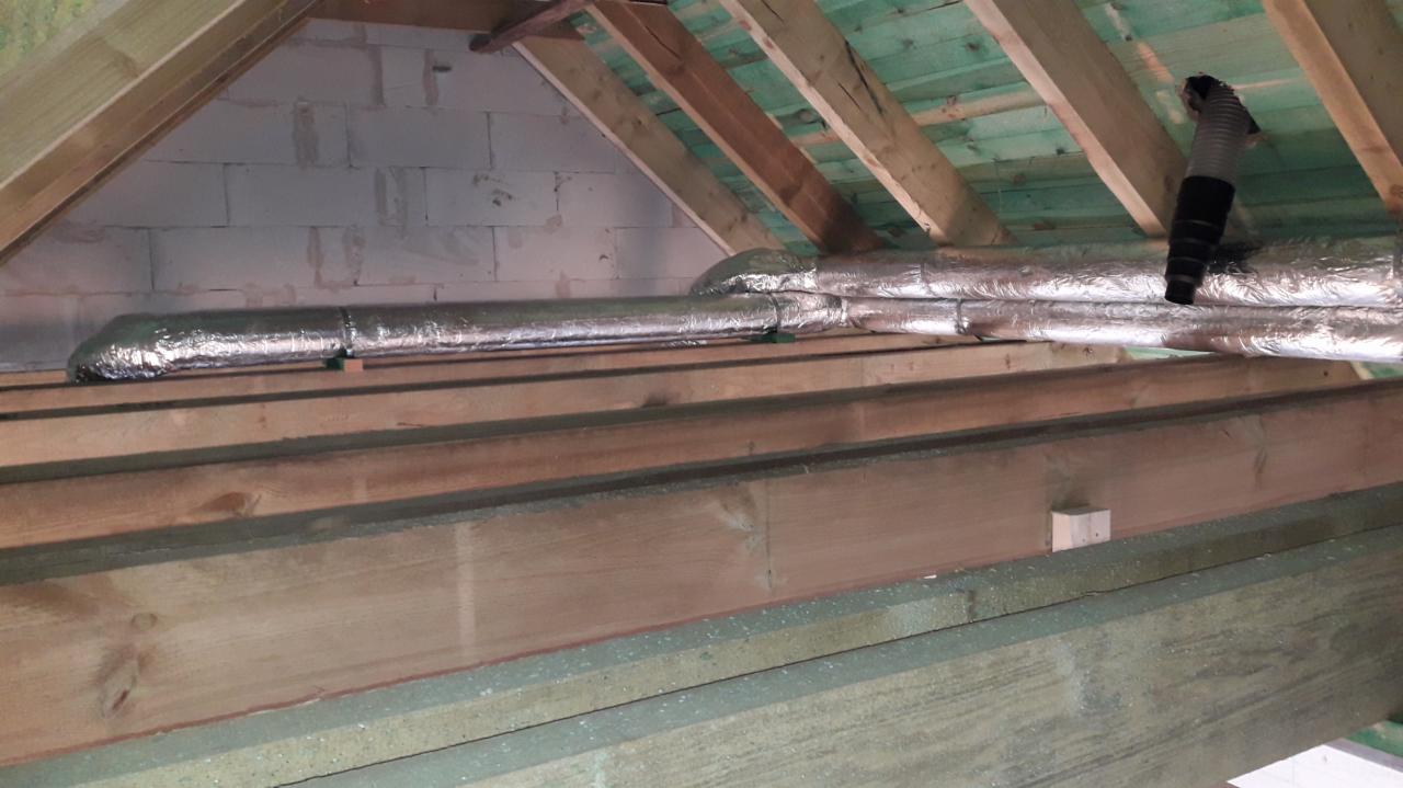 Izolowane przewody wentylacyjne w przestrzeni poddasza, widoczne drewniane elementy konstrukcyjne dachu i fragment ściany z bloczków.