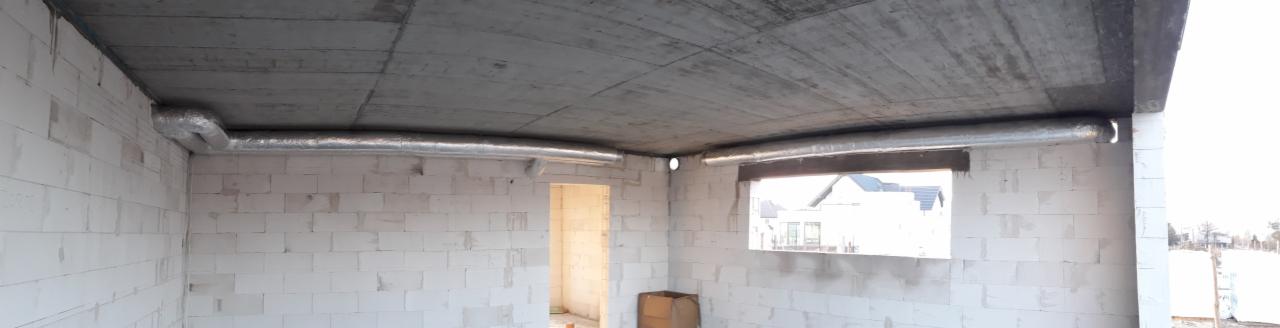 Wnętrze surowego budynku z widocznymi kanałami wentylacyjnymi poprowadzonymi pod betonowym stropem, ściany z białych bloczków, widok na zewnątrz przez otwór okienny.