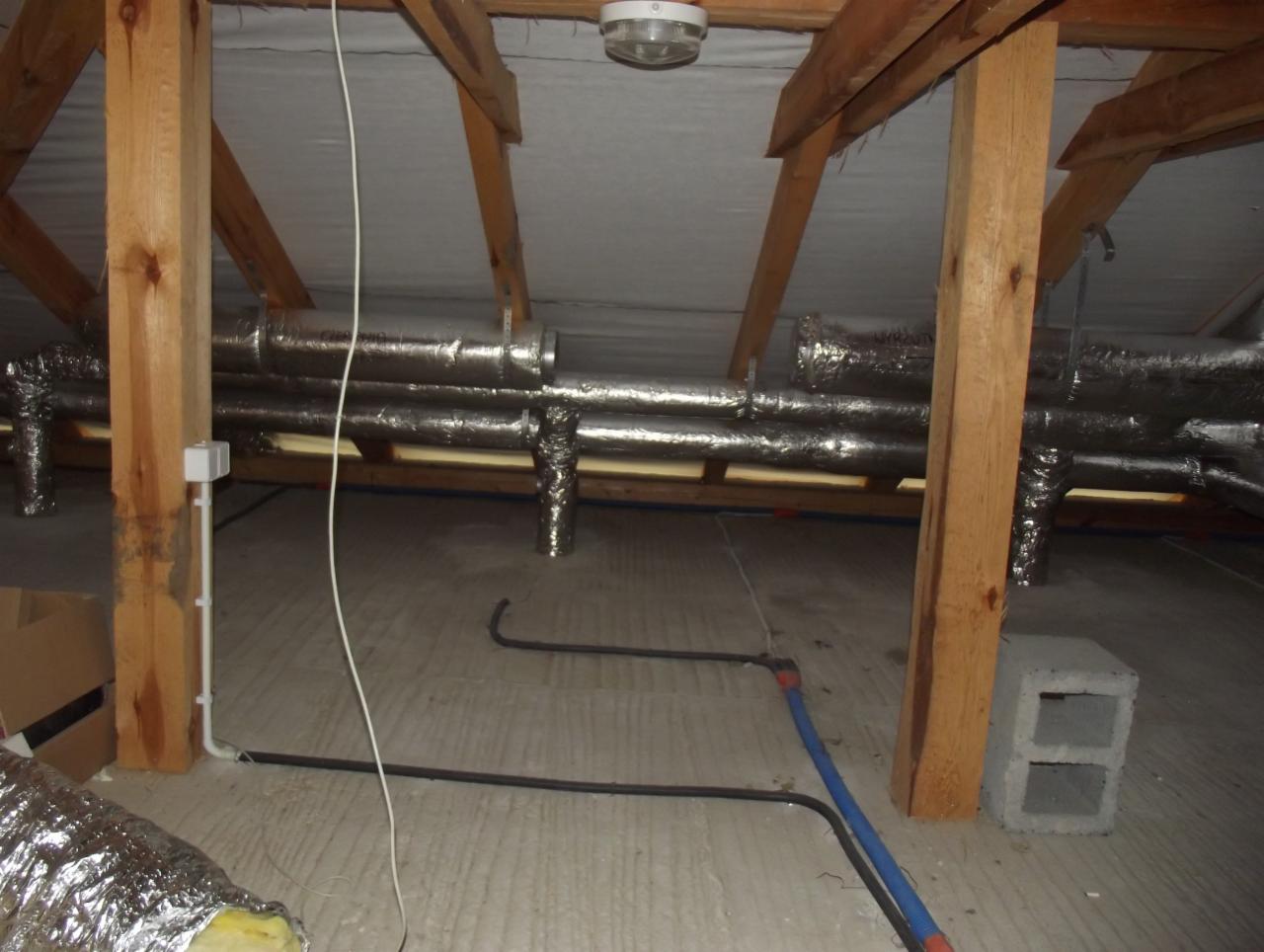 Instalacja przewodów wentylacyjnych w izolowanym poddaszu w Legionowie, widoczne drewniane elementy konstrukcyjne dachu i rury w srebrnej izolacji.