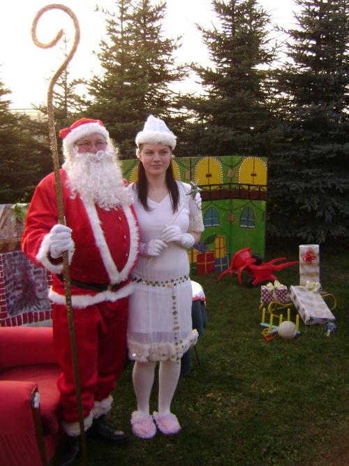 Mikołaj z laską i Śnieżynka pozują na tle scenografii z prezentami i choinkami.