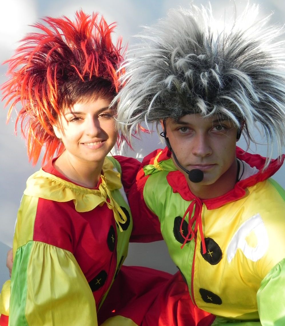 Dwoje klaunów w kolorowych kostiumach i perukach, uśmiechnięta kobieta z czerwoną peruką i mężczyzna z mikrofonem nagłownym oraz szaro-białą peruką.