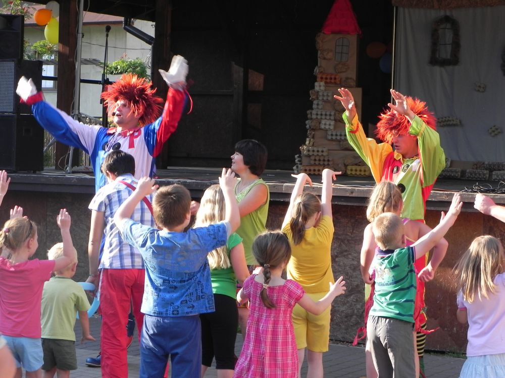 Dwóch klaunów w kolorowych kostiumach prowadzi zabawy z grupą dzieci na scenie podczas imprezy plenerowej; w tle dekoracja z kartonów po jajkach.