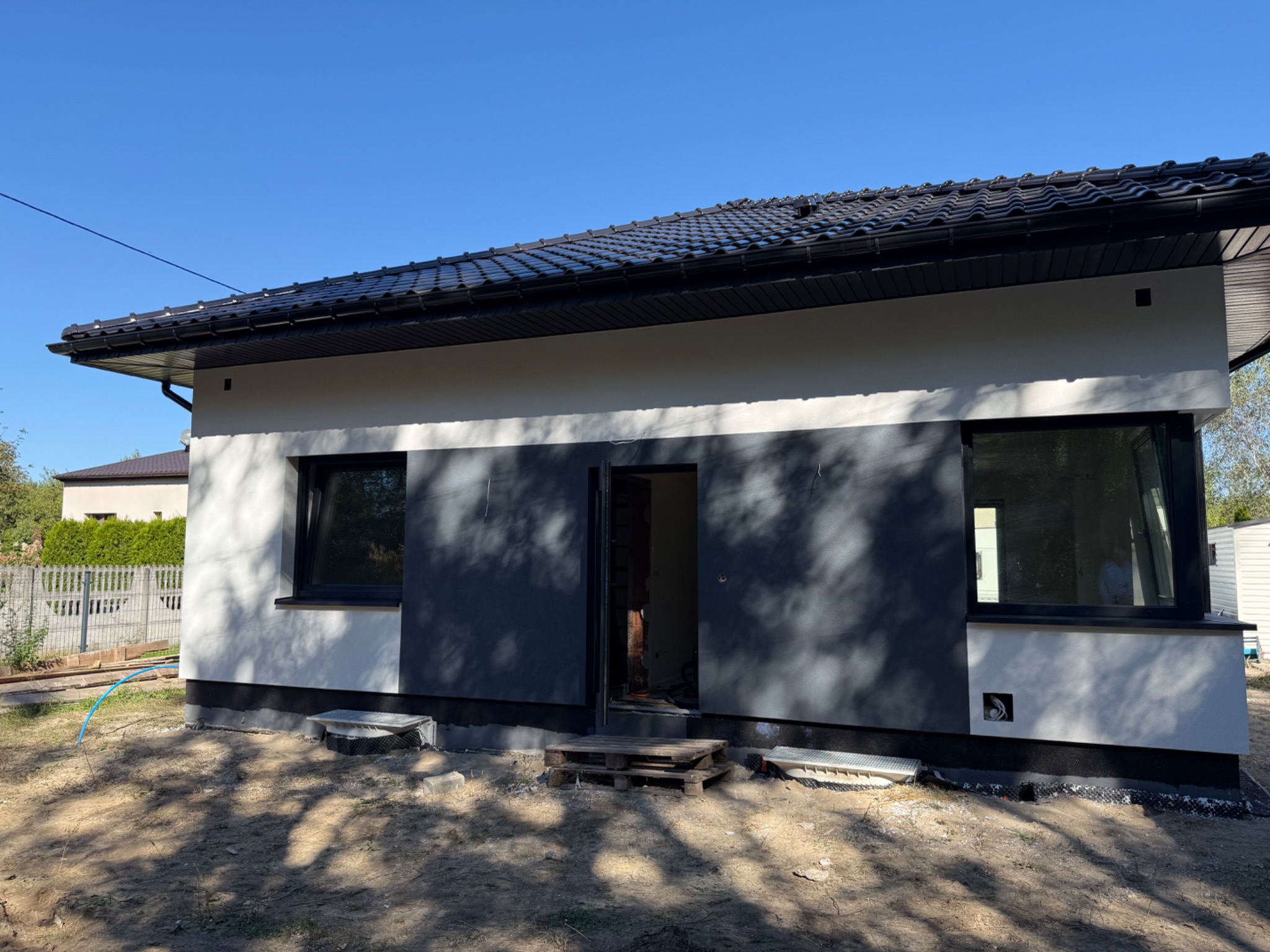 Budynek z częściowo wykończoną elewacją w odcieniach jasnej szarości i ciemnego grafitu, z czarnym dachem z dachówek i czarnymi oknami. Widoczne ocieplenie fundamentów.