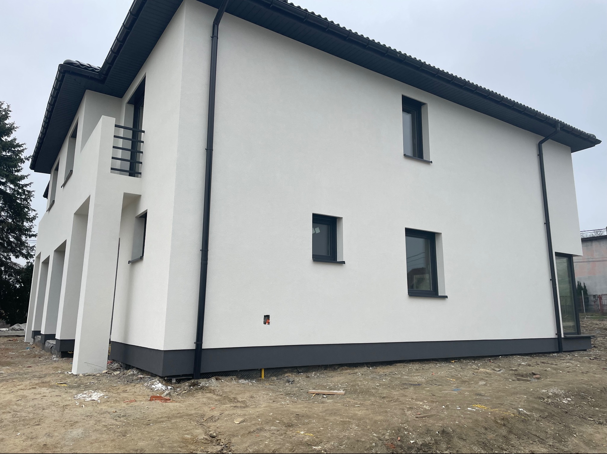 Nowoczesny dom jednorodzinny z białą elewacją i ciemnoszarym cokołem, widoczny z boku. Posiada czarne rynny, ciemne okna i balkon z metalową balustradą. Teren wokół budynku jest jeszcze...