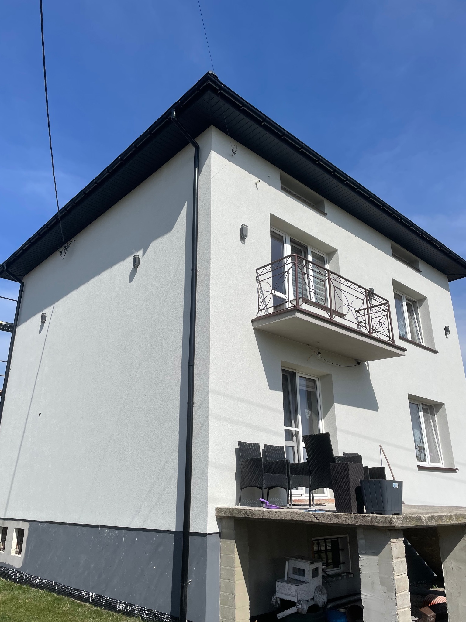 Jasnoszara elewacja dwupiętrowego domu z ciemnym cokołem, czarnym dachem i rynnami. Na piętrze balkon z kutą balustradą, na parterze taras z meblami ogrodowymi. Słoneczny dzień.