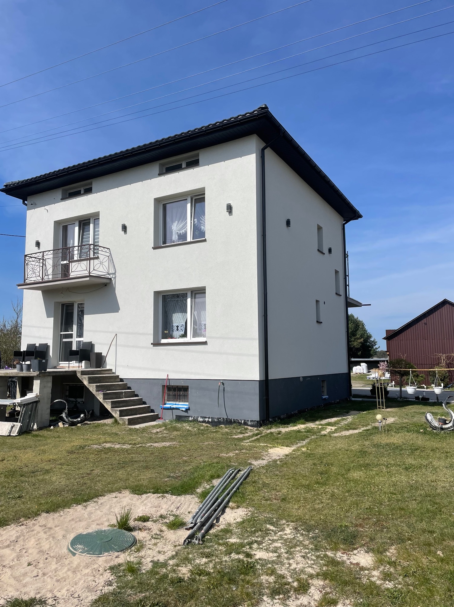 Nowo wykończony dwupiętrowy dom z białą fasadą, ciemnoszarym cokołem, czarnym dachem i ozdobnym balkonem. Widoczne betonowe schody i częściowo zagospodarowany teren.
