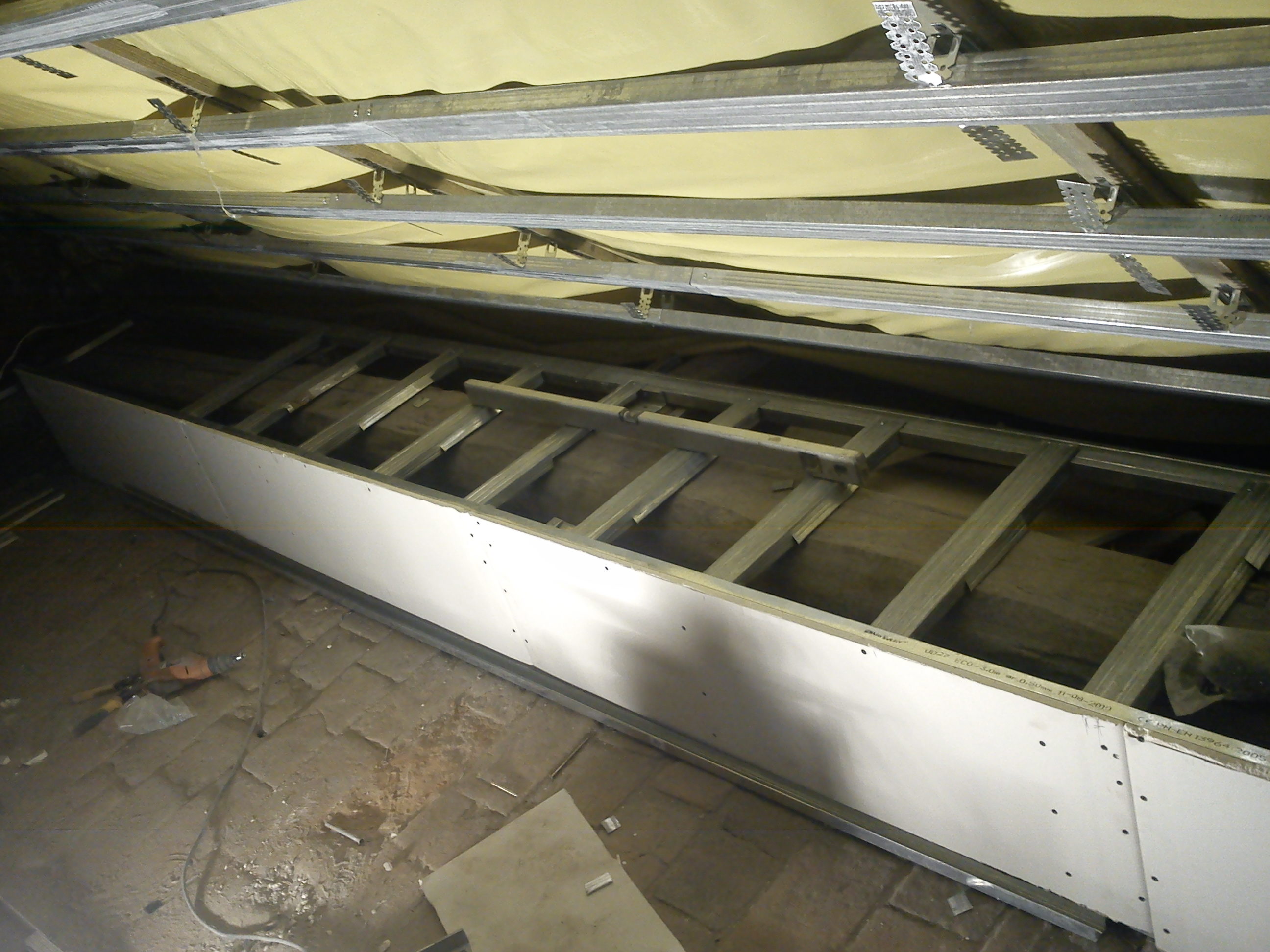 Szkielet sufitu podwieszanego z płyt kartonowo-gipsowych w trakcie montażu, widoczne profile metalowe, izolacja oraz narzędzia na podłodze.