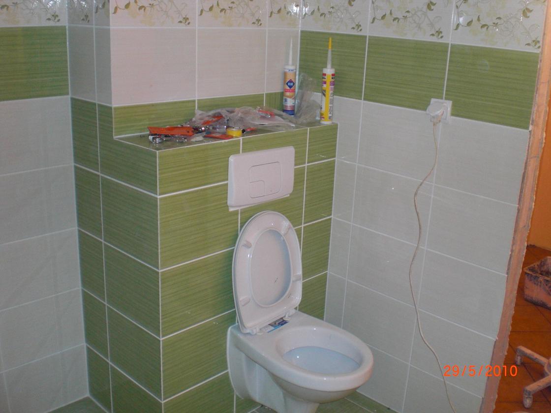 Wnętrze łazienki w trakcie remontu: widoczne płytki w odcieniach zieleni i bieli, częściowo ułożone, narzędzia i materiały budowlane na obudowie stelaża WC, oraz niedokończone wykończenie ściany.