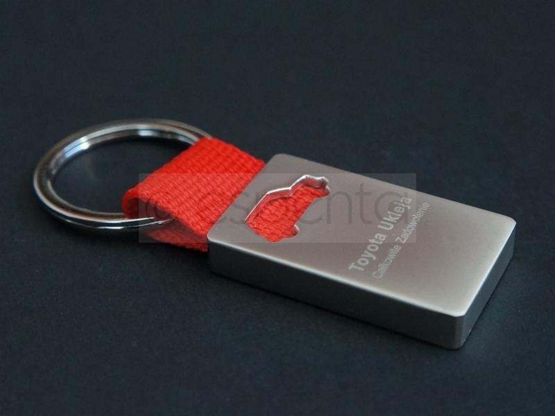 Metalowy brelok z logo Toyota Ukleja i wyciętym kształtem samochodu, zawieszony na czerwonym pasku i metalowym kółku, na ciemnym tle.