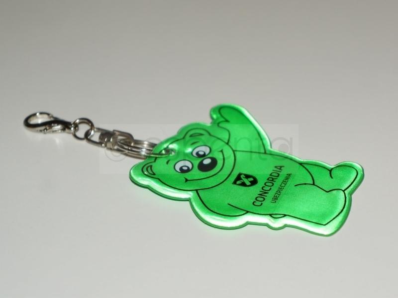 Brelok w kształcie zielonego misia z logo firmy ubezpieczeniowej Concordia, wykonany z odblaskowego materiału.