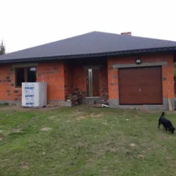 Niedokończony dom jednorodzinny z częściowo zamontowanymi oknami, brązowymi drzwiami wejściowymi i garażowymi, paleta bloczków Solbet przed oknem, pies na trawniku.