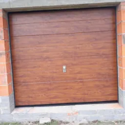 Brązowa, segmentowa brama garażowa z poziomymi panelami, zamontowana w surowym murze z czerwonej cegły, oświetlona zewnętrznym reflektorem LED.