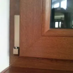 Fragment okna z brązową ramą imitującą drewno, widoczny czujnik otwarcia okna w kolorze kremowym, biała ściana w tle.