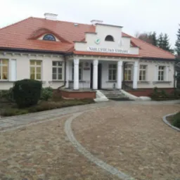 Budynek Nadleśnictwa Sarnaki z brukowanym podjazdem i chodnikiem z kostki. Jasna elewacja, czerwony dach. Ujęcie z poziomu terenu, pochmurne niebo.