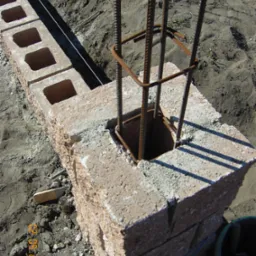 Wznoszenie ścian fundamentowych z bloczków betonowych z wkładką zbrojeniową, widoczna lina wyznaczająca prostą linię fundamentu.