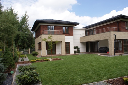 Dwukondygnacyjny dom z jasną elewacją i drewnianymi akcentami, widok z zadbanego trawnika z elementami małej architektury ogrodowej.