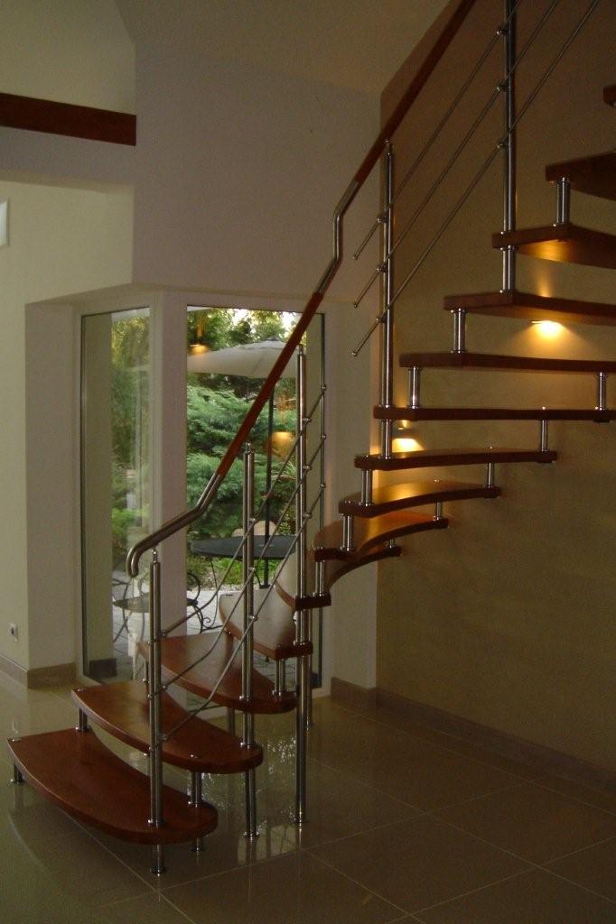 Spiralne schody z drewnianymi stopniami i nierdzewną balustradą, oświetlone punktowymi lampkami pod stopniami, widok z dołu na tle okna z widokiem na ogród.