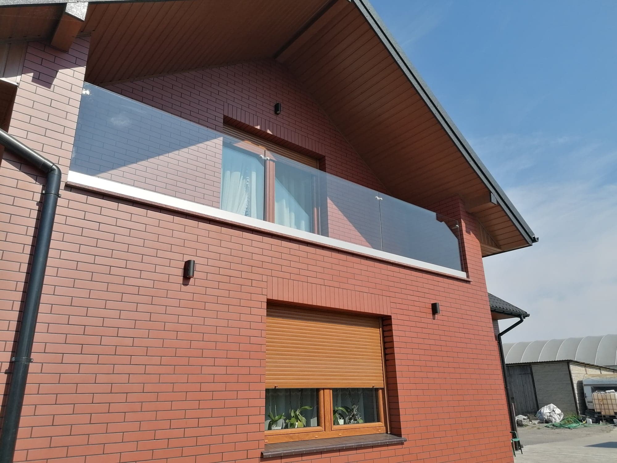 Nowoczesny balkon z balustradą ze szkła bezramowego, zamontowaną na jasnej, poziomej belce, na tle elewacji z cegły klinkierowej i błękitnego nieba.