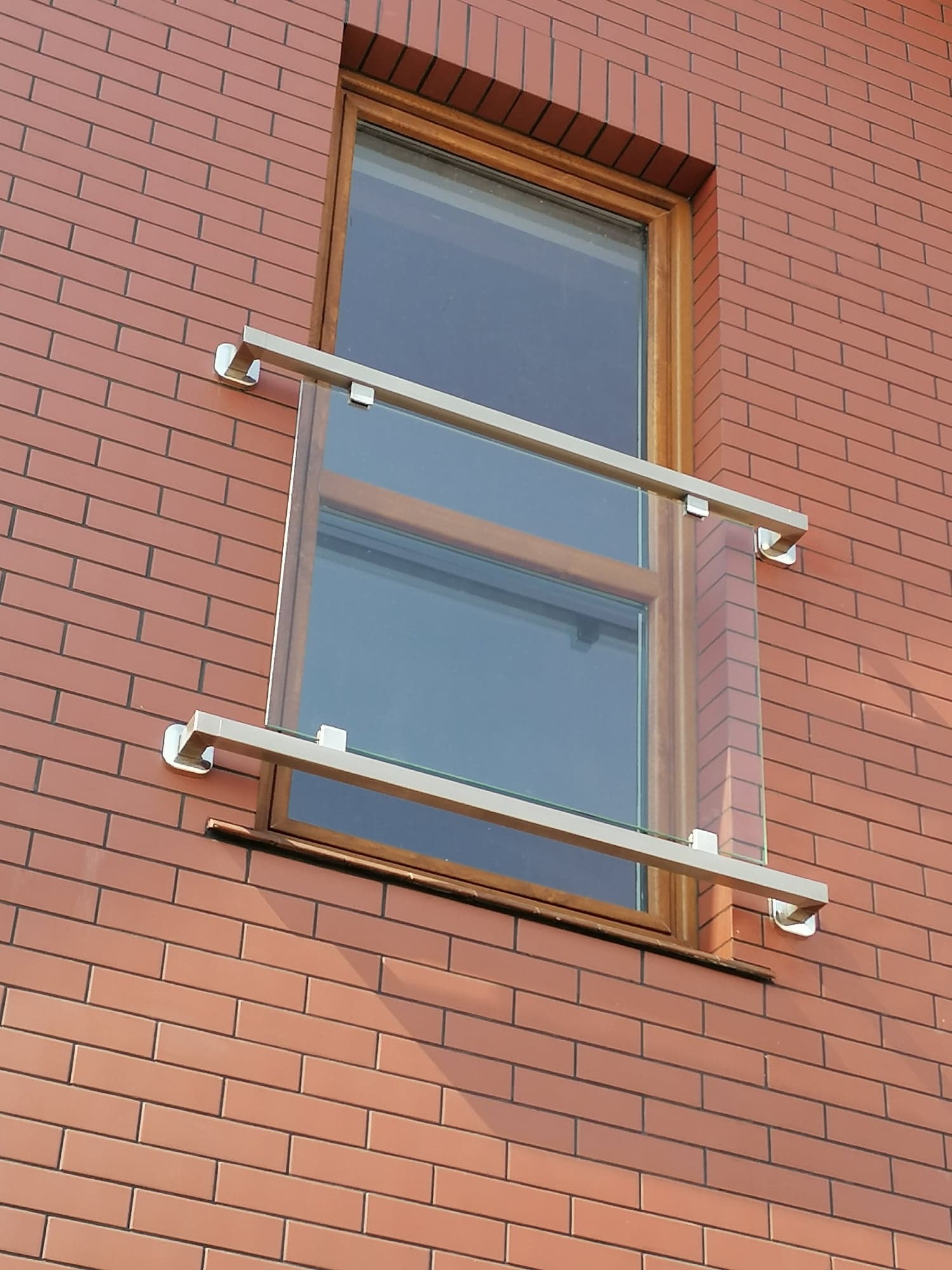 Szklana balustrada balkonowa z nierdzewnymi mocowaniami, zamontowana przy oknie na elewacji z cegły klinkierowej.