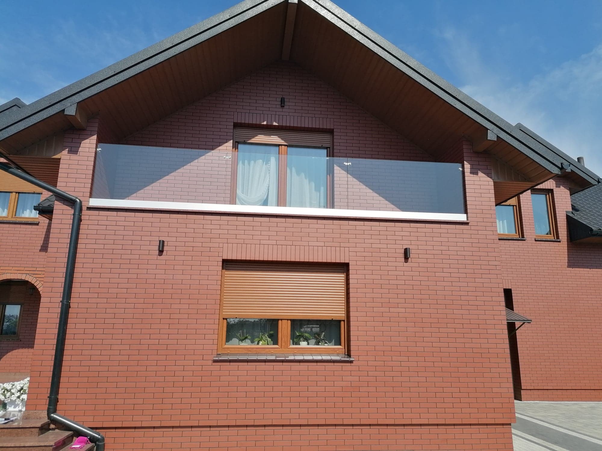 Nowoczesny dom z cegły z balkonem zabezpieczonym szklaną balustradą, okna z roletami zewnętrznymi, widok z dołu na elewację budynku.