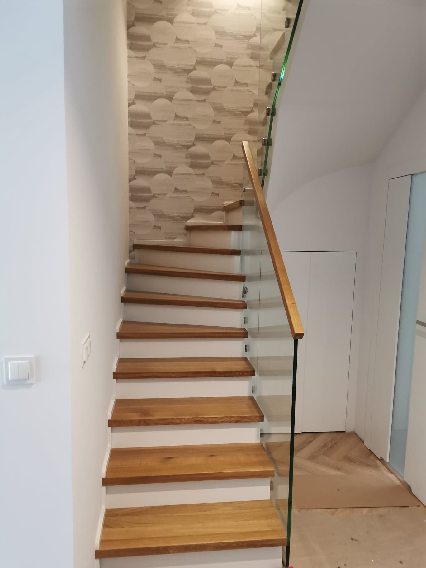 Wewnętrzne schody z drewnianymi stopniami i szklaną balustradą z drewnianą poręczą, wzorzysta tapeta w tle, widok od dołu.