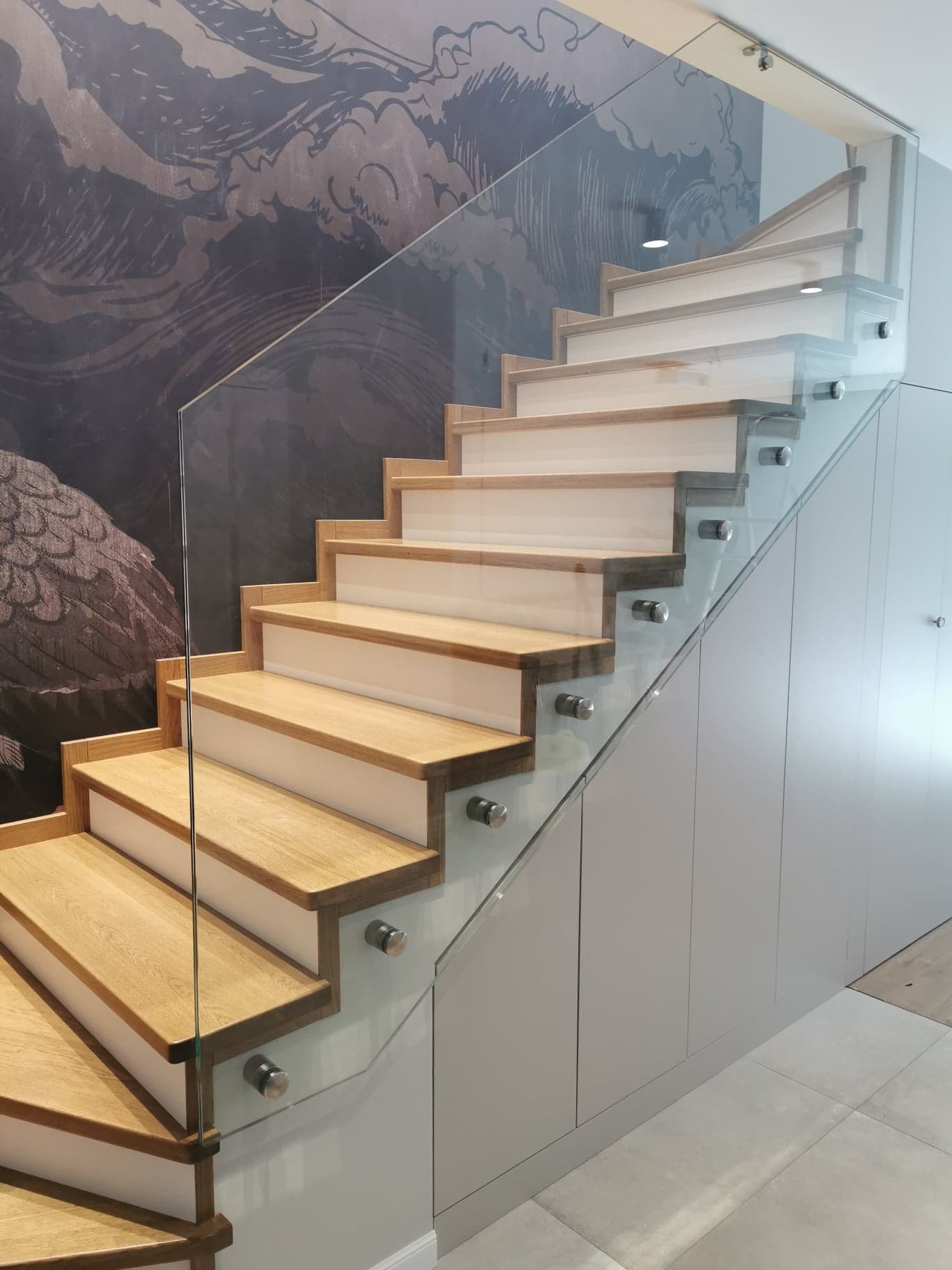 Schody drewniane z białymi podstopnicami i szklaną balustradą mocowaną na srebrnych uchwytach, zlokalizowane w Kaliszu, na tle ściany z artystyczną tapetą.