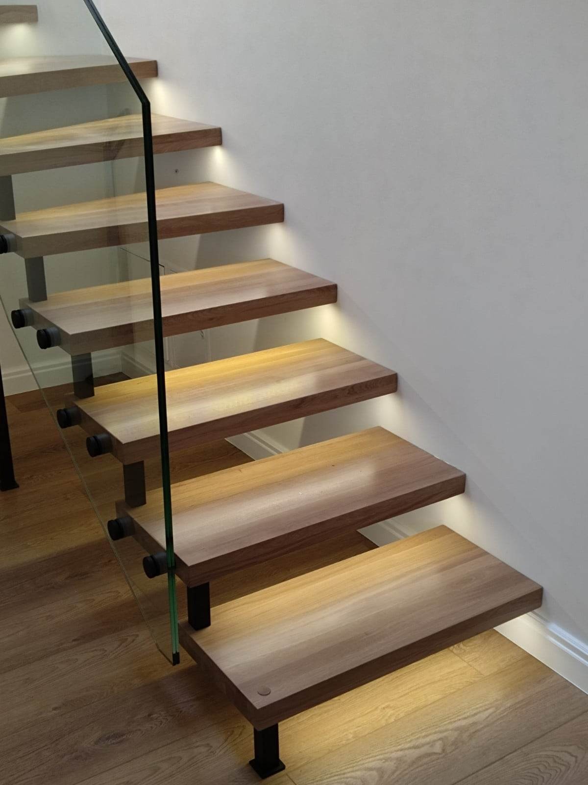 Nowoczesne schody z drewnianymi stopniami, podświetleniem LED, szklaną balustradą i czarnymi elementami mocującymi, widoczne z boku na tle jasnej ściany i drewnianej podłogi.