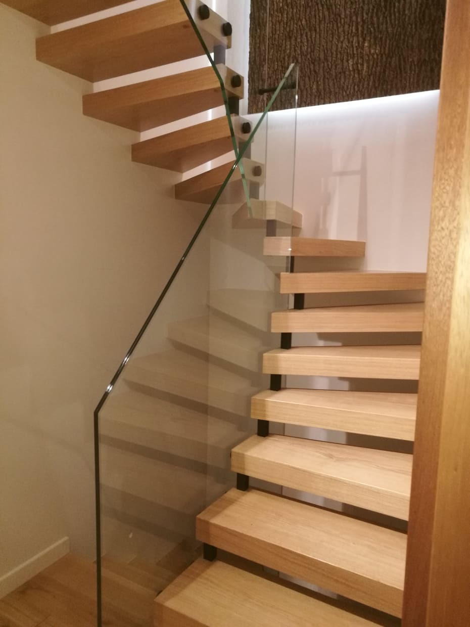 Lekkie, drewniane schody wspornikowe z czarnymi metalowymi łącznikami i szklaną balustradą, widok z dołu, oświetlenie LED w tle.