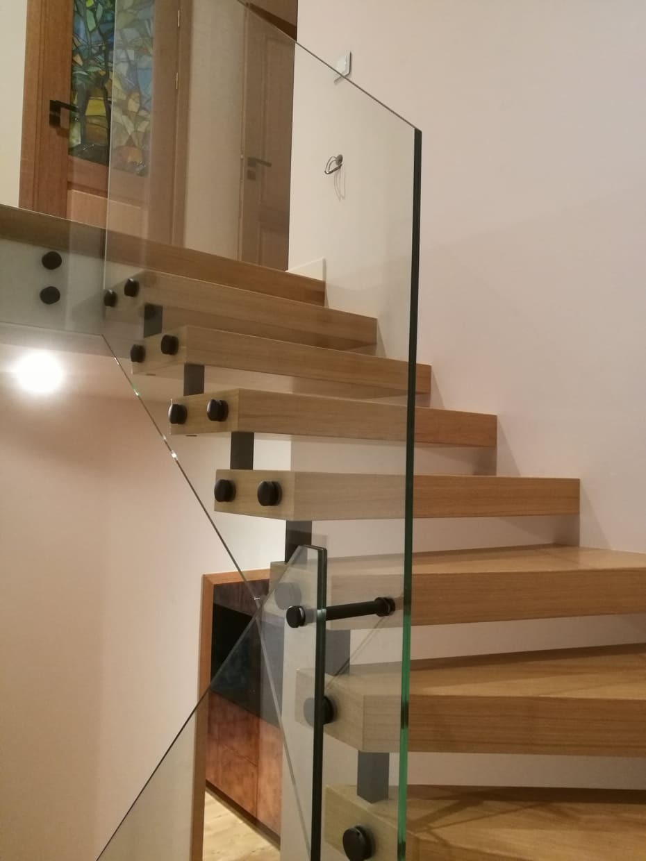 Nowoczesne schody z jasnego drewna z przezroczystą szklaną balustradą mocowaną czarnymi śrubami, widoczne drzwi z witrażem w tle.