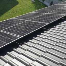 Rząd paneli słonecznych na szarym dachu z dachówki ceramicznej, widok z góry, w tle trawnik i ogrodzenie.