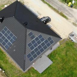 Szymel-bud - Widok z góry na dom z ciemnym dachem pokrytym panelami słonecznymi; obok zaparkowany samochód osobowy z drabiną na dachu i przyczepka z materiałami, sugerujące montaż lub serwis instalacji.