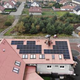 Szymel-bud - Widok z góry na dom z czerwoną dachówką, na której zamontowane są panele fotowoltaiczne; w tle zabudowa jednorodzinna i drzewa.