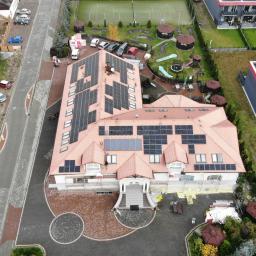 Szymel-bud - Widok z drona na duży budynek z zainstalowanymi panelami słonecznymi na dachu o różnym układzie i rozmiarach, obok budynki mieszkalne i teren rekreacyjny.