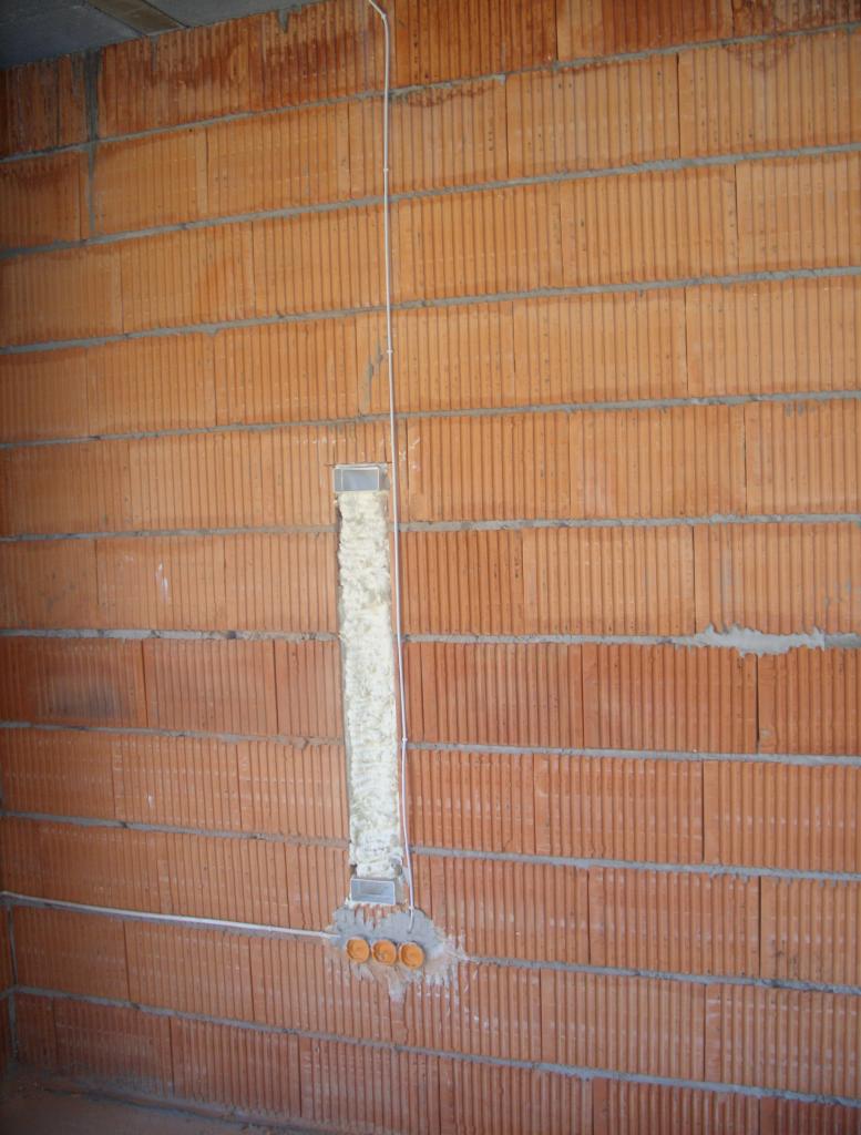 Instalacja elektryczna w ścianie z czerwonej cegły dziurawki, z puszkami i przewodem pionowym zabezpieczonym pianką montażową.