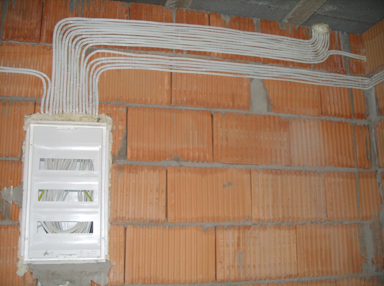 Rozprowadzona instalacja elektryczna w białych rurach, wewnątrz niezamkniętej skrzynki rozdzielczej, na tle ściany z czerwonej cegły dziurawki.