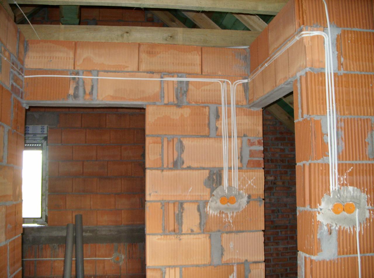 Wnętrze w trakcie budowy: ściany z czerwonej cegły, widoczne przewody elektryczne w białych rurkach, puszki instalacyjne. Surowy stan, instalacja elektryczna w trakcie montażu.