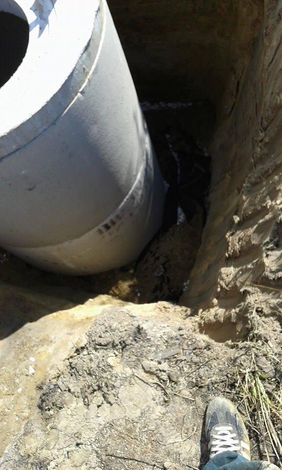 Betonowa rura w wykopie, widoczny fragment buta pracownika, ziemia i błoto.