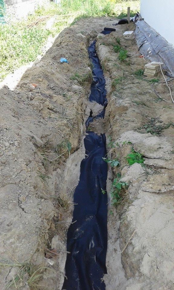 Wykop w ziemi wyłożony czarną geowłókniną, przygotowany pod instalację sanitarną, widoczne elementy instalacji na powierzchni.