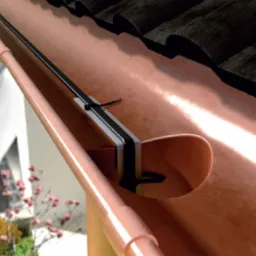 Instalacja przewodu grzejnego w rynnie dachowej z bliska, zabezpieczona uchwytem i opaską zaciskową, z widocznym fragmentem dachu z dachówki ceramicznej i elementami elewacji budynku w tle.
