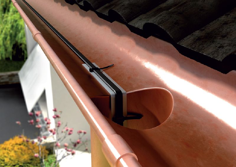 Instalacja przewodu grzejnego w rynnie dachowej z bliska, zabezpieczona uchwytem i opaską zaciskową, z widocznym fragmentem dachu z dachówki ceramicznej i elementami elewacji budynku w tle.