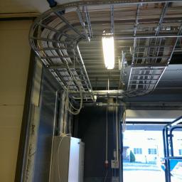 Artel Electric - Metalowe koryta kablowe pod sufitem w hali przemysłowej, widoczne przewody, oświetlenie LED, skrzynki elektryczne na ścianie, częściowo otwarte drzwi garażowe z widokiem na budynek na zewnątrz.
