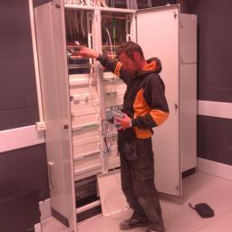 Artel Electric - Mężczyzna w roboczym ubraniu sprawdza połączenia w otwartej szafie sterowniczej z licznymi kablami i przewodami, używając specjalistycznego miernika.