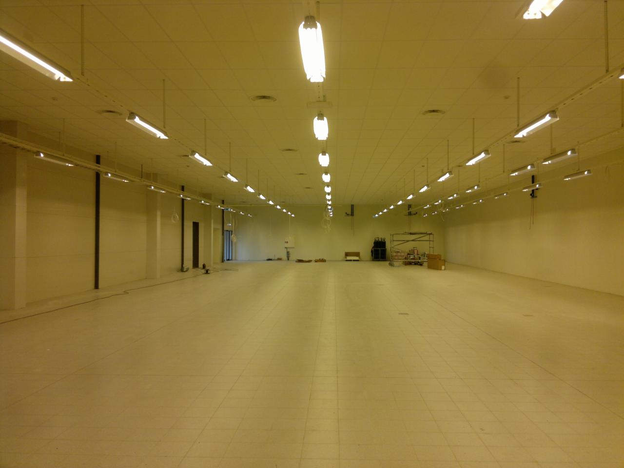 Przestronne wnętrze hali magazynowej z rzędem wiszących lamp fluorescencyjnych, widoczne instalacje elektryczne na suficie, ściany w odcieniach beżu, podłoga wyłożona płytkami, w tle widoczne...
