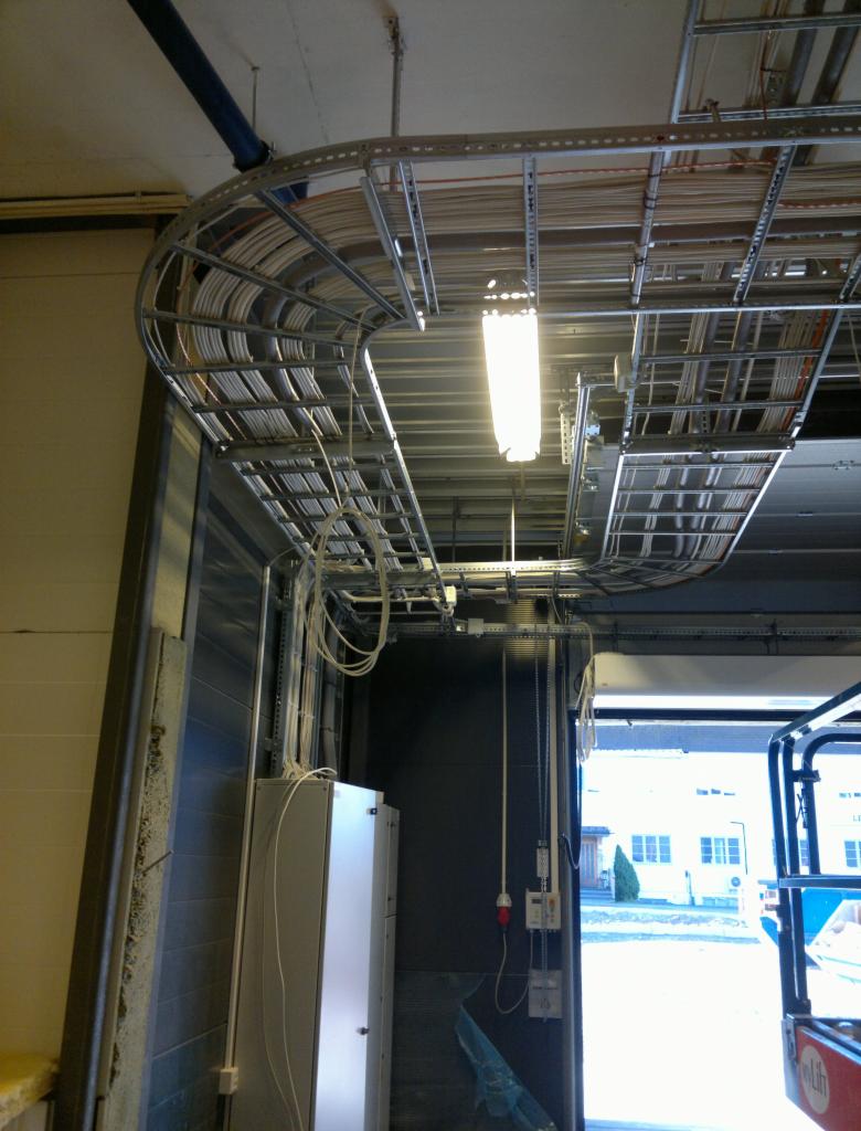 Metalowe koryta kablowe pod sufitem w hali przemysłowej, widoczne przewody, oświetlenie LED, skrzynki elektryczne na ścianie, częściowo otwarte drzwi garażowe z widokiem na budynek na zewnątrz.